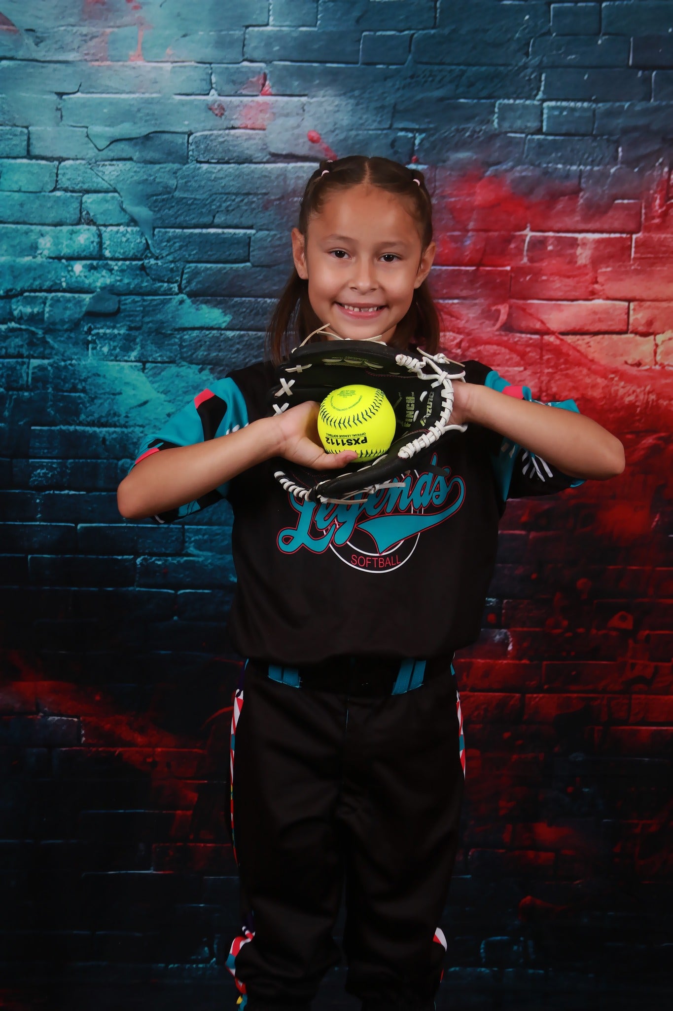 Girl holding a yellow baseball standing in front of a turquoise brick wall backdrop