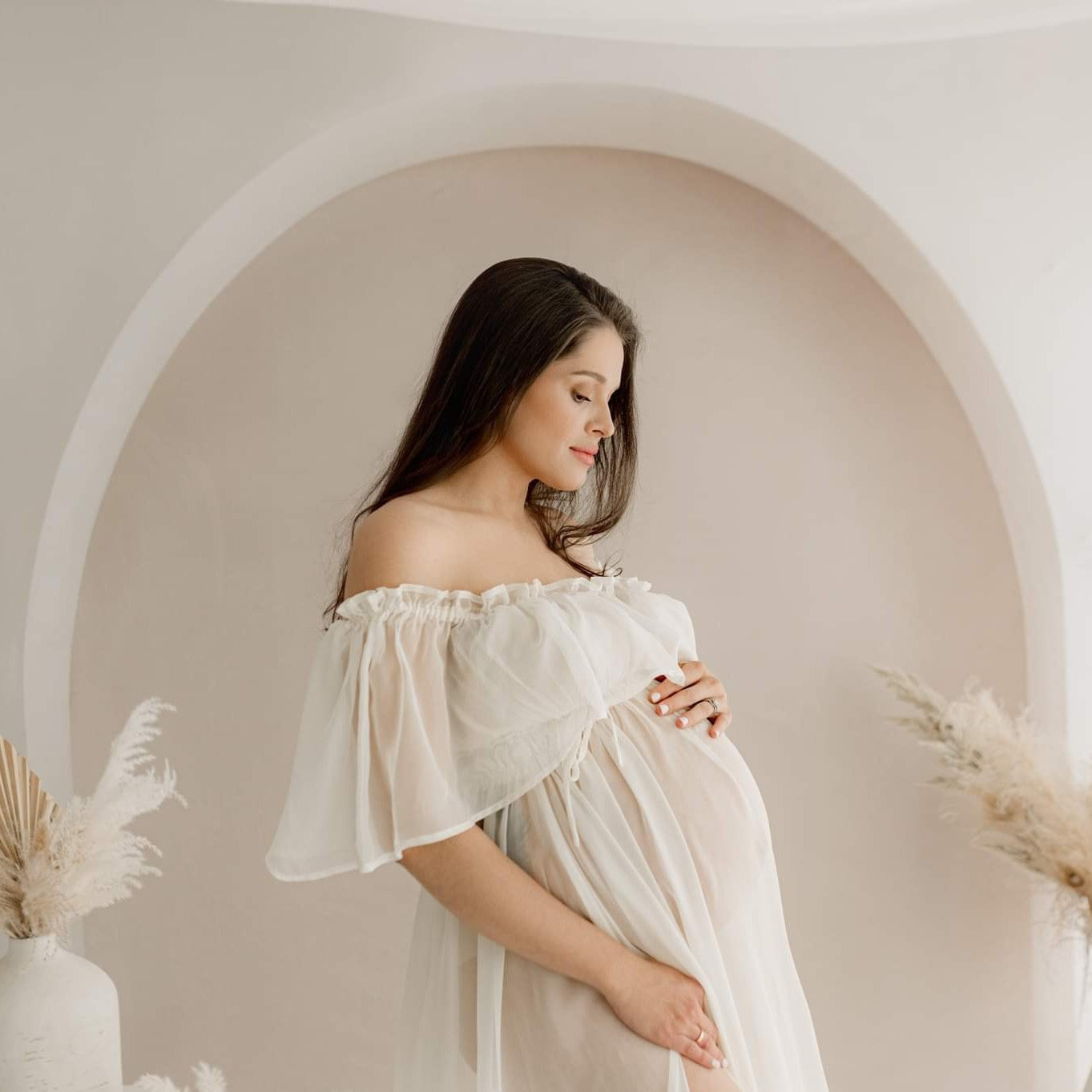 The expectant mother touching her pregnant belly in front of the boho simple white arch
